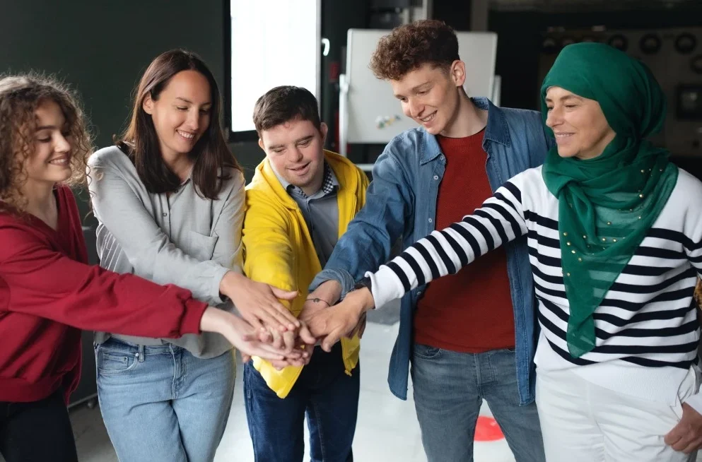 Multicultural friends all putting hands in the middle of a circle, one man disabled with down syndrome